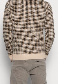 Beżowy sweter z dzianiny w czarno-brązowy wzór w kratę. Ribbed cuffs i dół, okrągły dekolt. Nosi się go na jasnoszarych dżinsach.