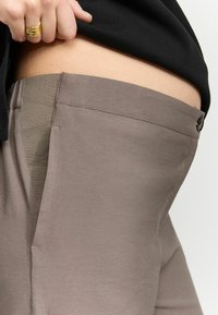 Lys taupe bukser med en glat tekstur og sidelomme, har en grå talje og en knaplukning. Sømmene er synlige.