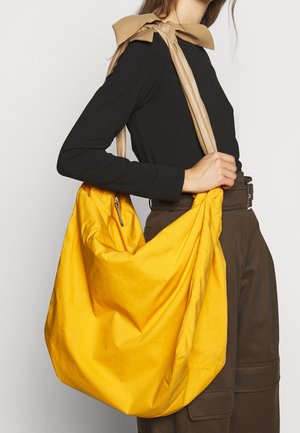 Gul tygkasse med en avslappnad design, med ett mjukt beige handtag. Inkluderar ett blixtlås och har en mjuk textur.