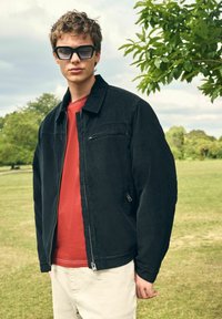 Zwarte corduroy jas met een ritstas, gecombineerd met een rood shirt en crèmekleurige broek. Het model draagt oversized zwarte zonnebrillen in een buitenomgeving.
