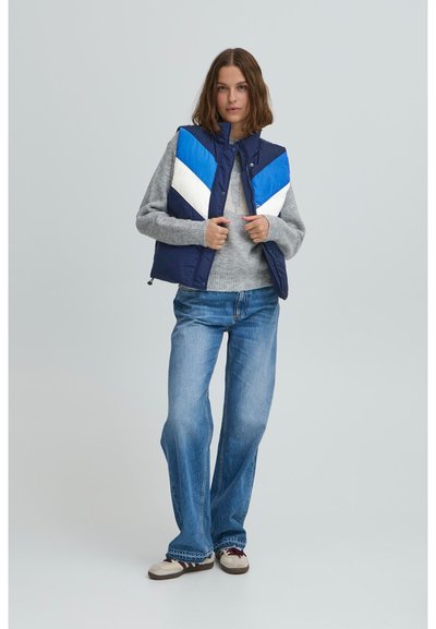 Dunkelblau gesteppte Weste mit blau-weißen Zickzackdetails, getragen über einem grauen Pullover, kombiniert mit blauen Jeans und beigen Schuhen.