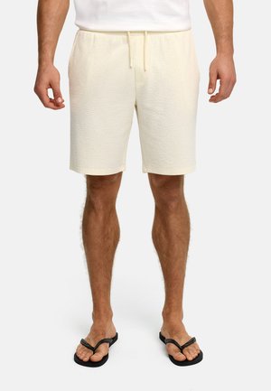 Mann trägt strukturierte, elfenbeinfarbene Shorts mit Kordelzug und schwarze Flip-Flops, steht vor einem schlichten weißen Hintergrund.