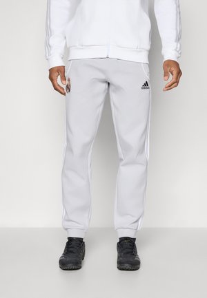 Homme portant un pantalon de survêtement blanc Adidas avec les logos de Real Madrid et d'Adidas, associé à des chaussures de sport noires, debout devant un fond uni.
