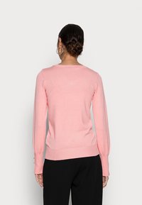 Femme avec des cheveux bruns en chignon, portant des boucles d'oreilles en argent, un pull rose à manches longues et un pantalon noir, se tenant de dos face au spectateur.