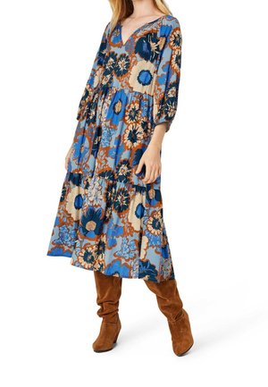 Robe fleurie bleu et beige avec un col en V, des manches trois-quarts et une jupe à volants. Associée à des bottes hautes en daim marron.