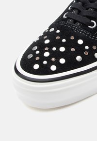 Adidași negri din suede cu ținte metalice și pietre prețioase, talpă din cauciuc alb cu o bandă neagră, design cu vârf rotund și șireturi negre.