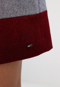 Falda de fieltro gris y burdeos con corte recto, que presenta una pequeña etiqueta metálica en el dobladillo inferior. Textura suave, sin patrones visibles.