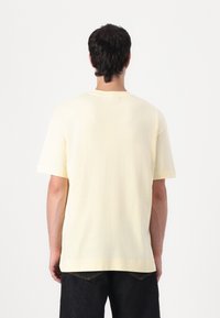 Light yellow cotton t-shirt, short sleeves, relaxed fit, rounded neckline, features a seam detail at the back. Worn with dark jeans.