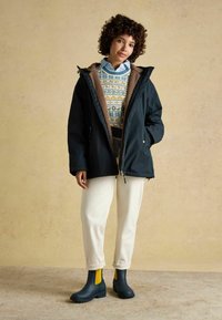 Čierna vonkajšia bunda s hnedým flísovým podkladom, cez vzorovaný sveter. Biele nohavice a tmavomodré dažďové topánky so žltými akcentmi.