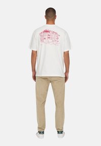 Weißes Baumwoll-T-Shirt mit einer roten Illustration eines Bodegas auf dem Rücken. Kurze Ärmel, lockere Passform, schlichte Vorderseite. Getragen mit hellbeigen Hosen.