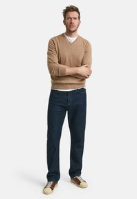 Brauner, gestrickter V-Ausschnitt-Pullover kombiniert mit dunkelblauen Jeans und braunen Sneakers mit beigefarbenen Sohlen. Das Outfit hat einen lässigen Schnitt und eine angenehme Textur.