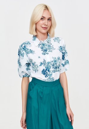 Kwiatowa bluzka z krótkim rękawem w niebieskie i zielone wzory na białym tle, zestawiona z spodniami w kolorze turkusowym o wysokim stanie.