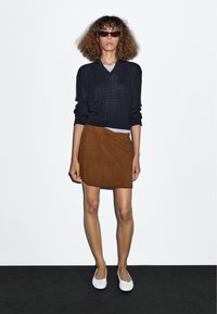 Schwarzer Strickpullover, brauner Wildlederrock und weiße Ballerinas. Präsentiert vor einfachem Hintergrund, der Textur- und Farbkontraste betont.