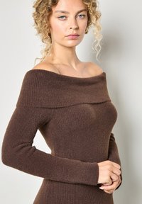 Bruine off-the-shoulder gebreide jurk met ribbeltextuur, lange mouwen en een aansluitende silhoeet, gemaakt van zachte, rekbare stof.