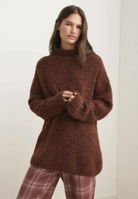 Frau mit langen braunen Haaren trägt einen übergroßen, flauschigen braunen Pullover und rosa karierte Hose, steht vor einem hellen neutralen Hintergrund.