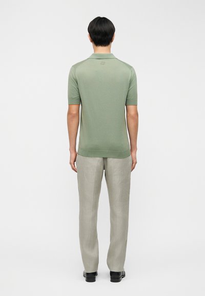 Πράσινο πόλο με στενή εφαρμογή και κοντά μανίκια, συνδυασμένο με ανοιχτό γκρι παντελόνι. Το πουκάμισο έχει ένα διακριτικό λογότυπο στο πίσω μέρος.