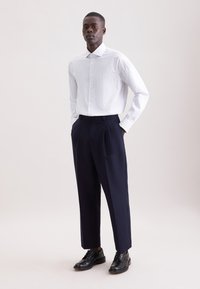 Camicia bianca a righe abbinata a pantaloni plissettati blu scuro e scarpe in pelle nere, con colletto classico e vestibilità su misura.