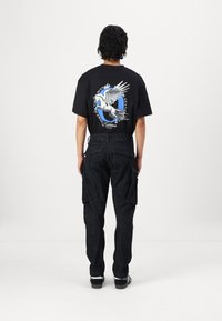 Black graphic t-shirt with winged horse design, paired with dark cargo pants featuring side pockets. Model faces away, showcasing clothing details.