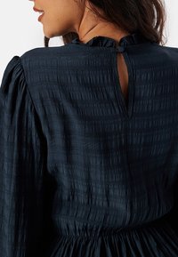 Robe bleu marine avec un tissu texturé, un col à volants et un dos en trou de clé. La taille froncée met en valeur la silhouette.