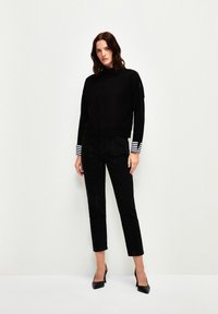 Schwarzer Pullover mit hohem Kragen und gestreiften Bündchen, kombiniert mit schwarzen geraden Hosen und spitzen schwarzen Schuhen, vor einem neutralen Hintergrund.