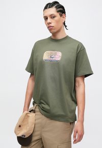 Olívazöld pamut póló retro stílusú rózsaszín és kék logóval az elején. Bő szabású, kerek nyakkivágással. Színes sapka a nadrágon pihen.