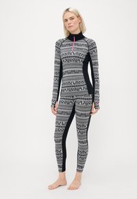 Nainen, joka on pukeutunut mustavalkoiseen kuvioituun pitkähihaiseen puseroon ja leggingseihin, joissa on peukalon reiät, seisoo paljain jaloin yksinkertaista taustaa vasten.