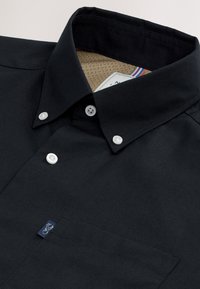 Black button-up shirt with white buttons, button-down collar, beige inner lining, and a small stitched logo on the chest pocket.
