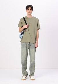 Olive green cotton t-shirt, casual fit, paired with light blue bag. Light wash denim jeans with a straight cut, white sneakers.