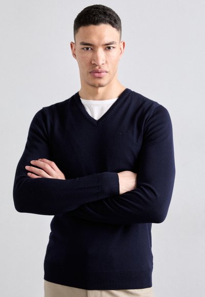 Mørkeblå V-hals sweater, strikket tekstur, lange ærmer, ribbede manchetter og kant, båret over en hvid skjorte. Modellen står med armene kors.