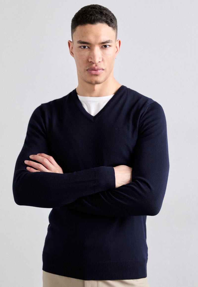 Pull-over en V bleu marine, texture tricotée, manches longues, bords côte aux poignets et à l'ourlet, porté sur une chemise blanche. Le mannequin se tient avec les bras croisés.