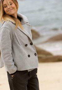 Grau melierte Wollmischung-Peacoat mit doppelter Knopfleiste, großen schwarzen Knöpfen und seitlichen Taschen, kombiniert mit dunklen Jeans vor einer Strandkulisse.