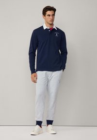 Mornarska polo majica dugih rukava s bijelim ovratnikom, u kombinaciji sa sivim trenirakama koje imaju mornarske manžete i logo. Model stoji u ležernim cipelama.