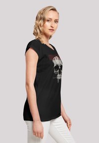 T-shirt noir avec un graphisme de crâne sur le devant, à manches courtes et coupe décontractée, porté avec un pantalon blanc.
