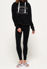Person som bär en svart Superdry-hoodie och svarta leggings, med vita Superdry-slidesandaler, stående mot en enfärgad bakgrund.