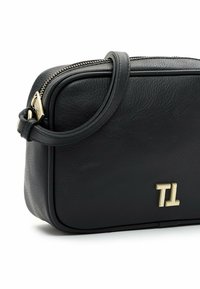 Sac bandoulière en cuir noir avec fermeture éclair dorée et initiales du logo "TL" à l'avant, équipé d'une sangle ajustable.