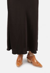 Mule in pelle marrone con punte appuntite e decorazioni ritagliate, abbinate a un vestito midi nero fluido con orlo scallop.
