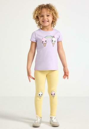Unge barn smiler, iført en lavendelfarvet t-shirt og gule leggings med tegneserieisvafler og et regnbuegrafik.