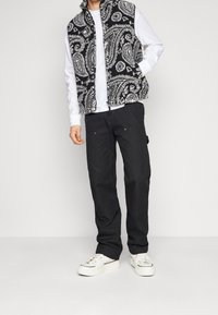 Gilet paisley bianco e nero con colletto alto, indossato sopra una camicia bianca a maniche lunghe. Pantaloni scuri e sneakers bianche completano l'outfit.