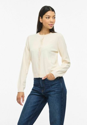 Frau trägt eine cremefarbene, geknöpfte Strickjacke und blaue Jeans, steht mit einer Hand in der Tasche und schaut nach links.