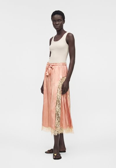 Γυναίκα με αμάνικο μπεζ τοπ, ροζ σατέν μίντι φούστα με δαντέλα και μαύρα σανδάλια χωρίς λουράκια με χρυσές καρδιές.