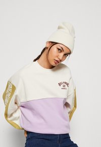 Bluza Billabong w kolorze kremowym, lawendowym i musztardowym, z blokami kolorów. Posiada logo na klatce piersiowej oraz wzorzyste akcenty na rękawach. Mieszanka bawełny.