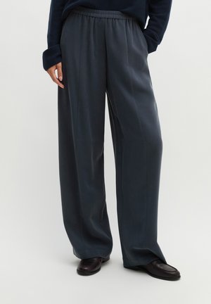 Personne portant un pantalon large sombre avec une taille élastique et des chaussures noires, debout avec une main dans la poche.