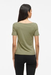 Olivgrön kortärmad topp med vid halsringning, mjuk textur och figurnära design. I kombination med svarta jeans. Bakvy visas.