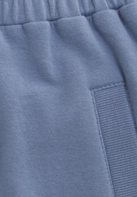 Lyseblå fleece joggebukser med elastisk linning, teksturert stoff og en sydd lappdetalj på siden. Glatt og myk tekstur.