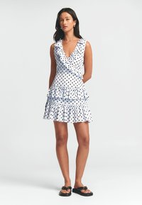 Valge kleit, millel on mustad täpid, volangi kaelus ja kihiline seelik. Kangas näeb välja kerge ja voolava disainiga.