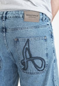 Jean denim bleu clair avec une poche arrière texturée comportant un design en broderie noire en relief et une étiquette de taille en cuir marron.