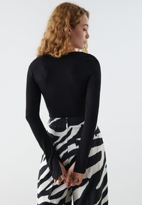 Zwart langemouwtop in combinatie met hoge taillebroek met een opvallend zwart-wit zebra-strippatroon. Model poseert met de handen achter de rug.