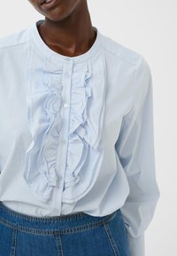 Donna che indossa una camicetta blu chiaro con bottoni e ruffles sul davanti, infilata in jeans di denim blu a vita alta.