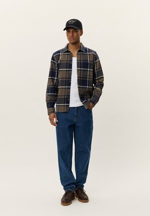 Chemise en flanelle à carreaux marron et marine, t-shirt blanc en dessous, jeans bleus et chaussures marron foncé. Porte une casquette noire avec un logo.