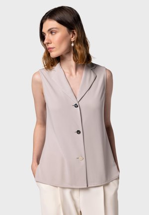 Gilet senza maniche di un leggero beige con colletto a punte, tre bottoni (due neri e uno crema) e una texture liscia. Abbinato a pantaloni crema.